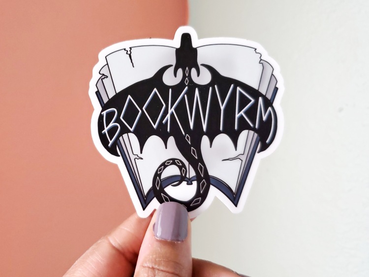 Sticker of a dragon silhouette flying over an open book. Over the wings of the dragon is the word "bookwyrm" written in rune-like letters. 