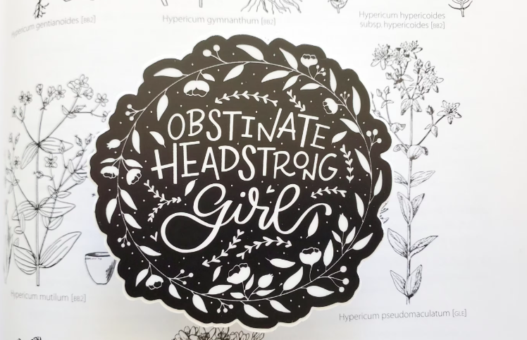 Black and white circle sticker: "Obstinate Headstrong Girl" in handwritten letters in the middle and a wreath of flowers all around. White letters and flowers with a black background. 