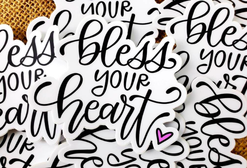 Die-cut sticker: Black letters on a white background. Letters are in modern calligraphy style, stating "bless your heart" with a pink heart as the period at the end of the sentence. 